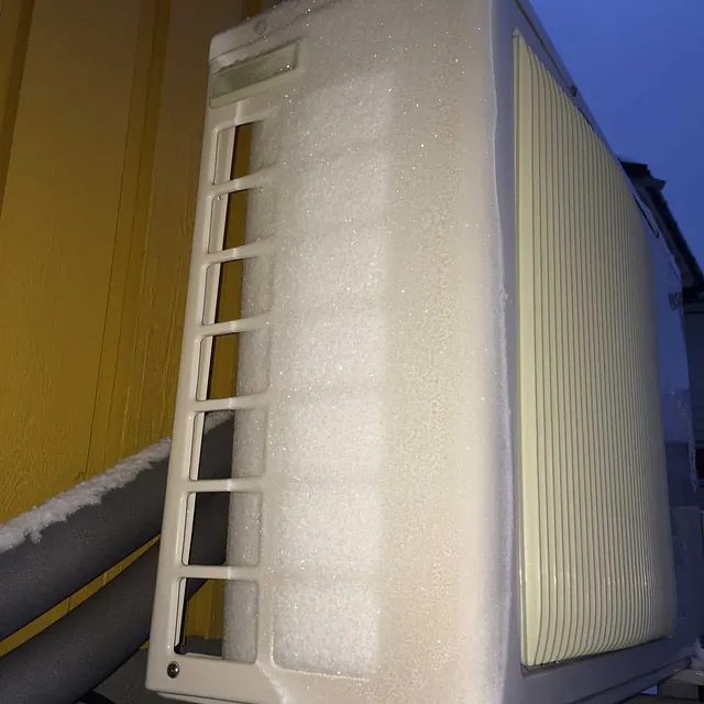 Varmepumpe utedel dekket av rim og snø, montert på vegg i kaldt vintervær.